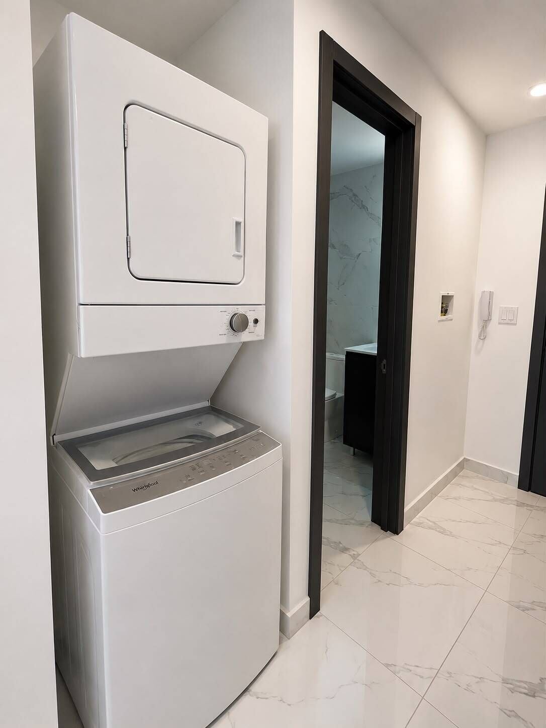 A compact laundry area with a front-loading washer and dryer, a door leading to a bathroom, and a tiled floor.