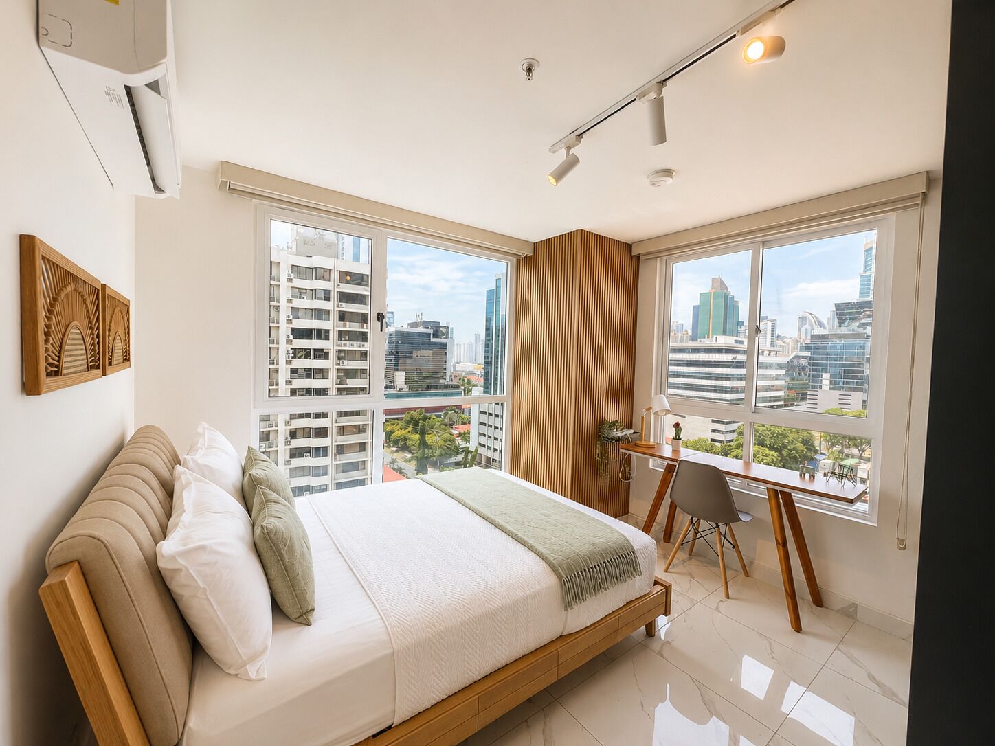 A modern bedroom with a large bed, a desk, and a view of the cityscape through large windows.