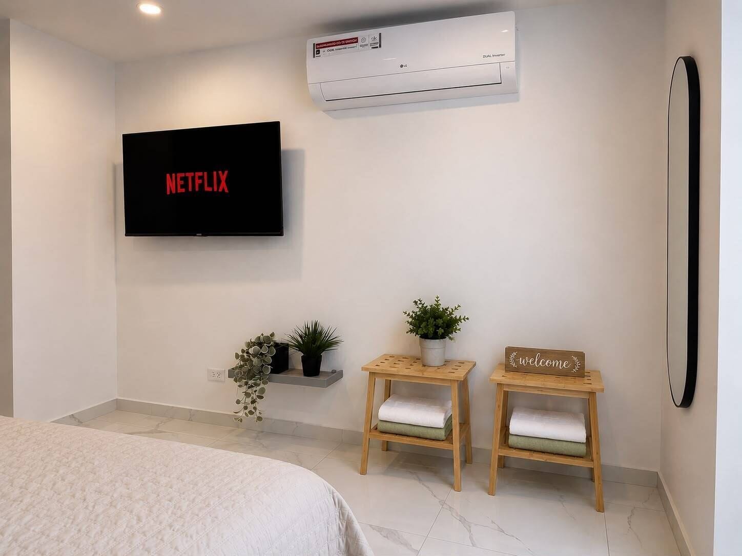 A modern hotel room with a flat-screen TV, an air conditioning unit, a bed, two wooden side tables with cushions, a welcome sign, and a potted plant.