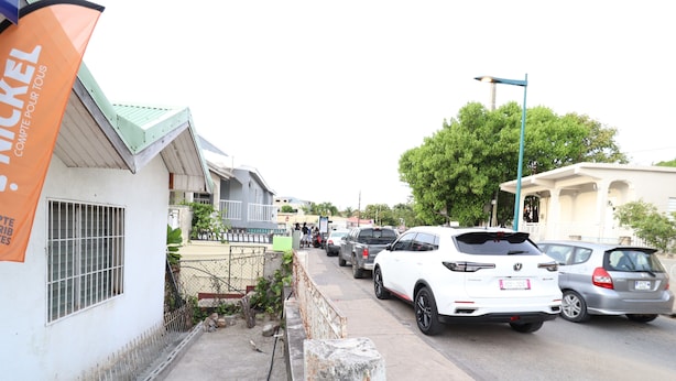 Rua com carros estacionados, um edifício com telhado verde e uma faixa com texto.