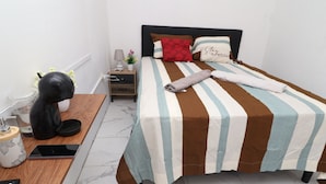 Uma cama bem arrumada com roupa de cama listrada, uma mesinha de cabeceira com um abajur e uma pequena planta, e uma cafeteira no chão.