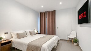 Ein modernes Schlafzimmer mit einem Bett, Nachttisch, Beistelltisch und einem an der Wand montierten Fernseher, der das Netflix-Logo zeigt.