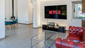 Ein modernes Wohnzimmer mit einem Flachbildfernseher, der 'NETFLIX' anzeigt, einem Glastisch, einem roten Ledersofa und einem Essbereich mit blauen Stühlen.
