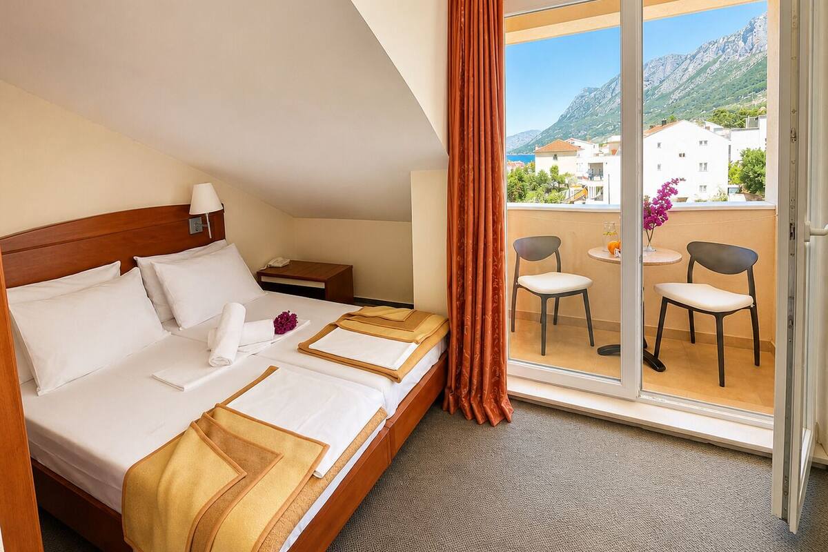 A hotel room with a bed, a nightstand, a window with curtains, and a balcony with chairs and a table.