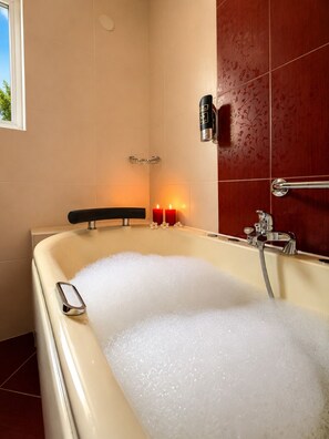 Eine Badewanne voller Blasen, eine Duschbrause zum Halten und zwei brennende Kerzen an der Wand.