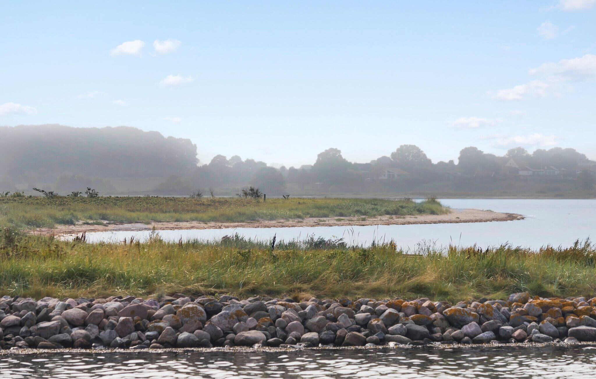 A calm lake with a rocky shoreline, surrounded by grassy areas and trees in the background.