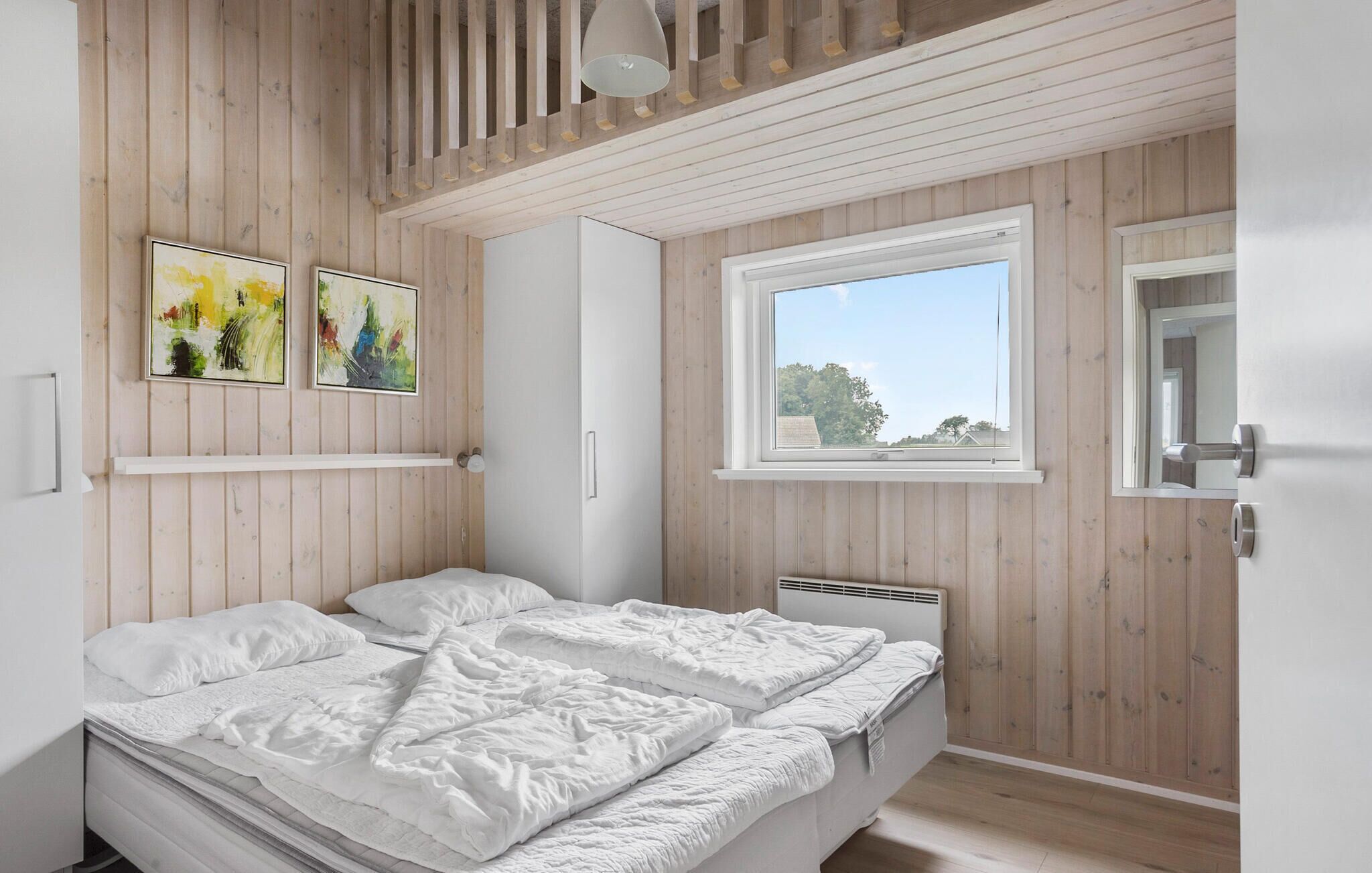 Ein Schlafzimmer mit Bett, Fenster, Holzwänden und zwei abstrakten Gemälden.