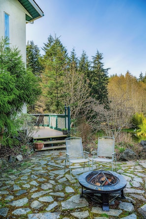 A stone patio with a fire pit, two chairs, and a wooden deck with a railing.