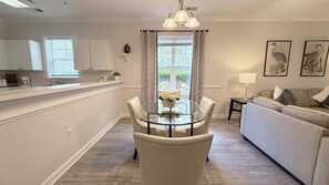 Eine moderne Küche mit weißen Schränken, eine Essecke mit Glastisch und Stühlen sowie ein Wohnzimmer mit Sofa und Lampe.