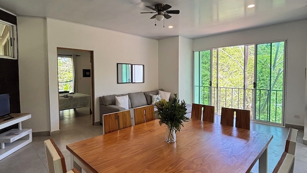 Un vaste espace dédié à la salle à manger, avec une grande table en bois, des chaises modernes, un ventilateur de plafond et de grandes fenêtres offrant une vue sur la verdure.