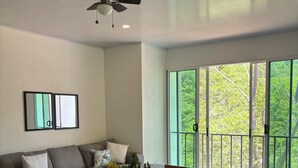 Un espace repas moderne avec une table en bois, des chaises, un ventilateur de plafond et une grande fenêtre donnant sur un paysage verdoyant.