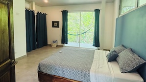 Une chambre à coucher avec un lit, un ventilateur de plafond, une fenêtre avec des rideaux et une vue sur les arbres à l’extérieur.