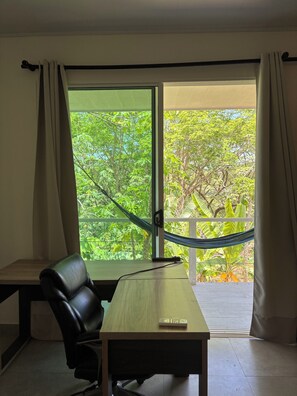 Ein Zimmer mit Schreibtisch, Stuhl und Blick auf Grün durch ein großes Fenster.