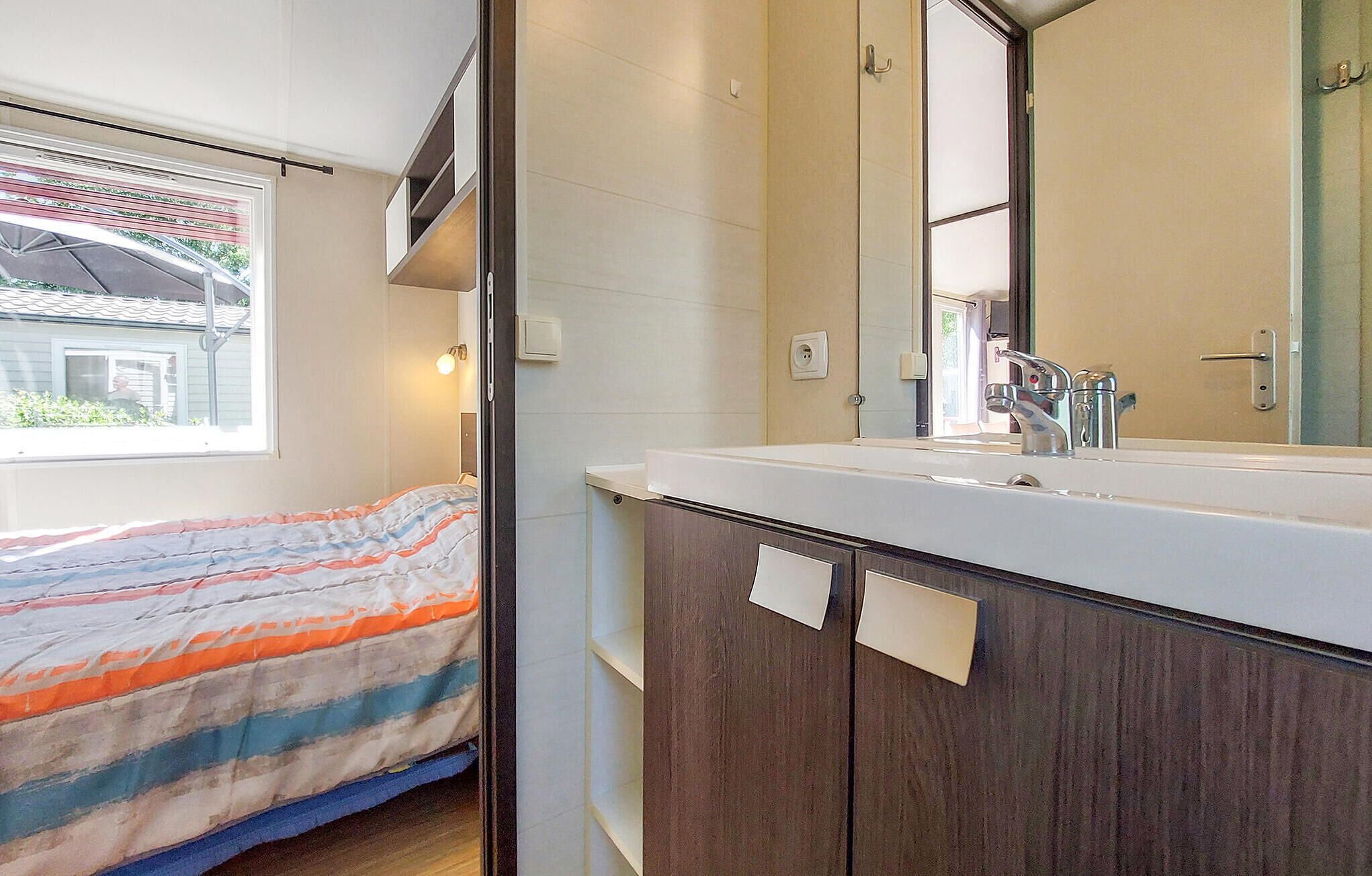 A bedroom with a bed, a window, a bathroom with a sink and mirror, and a door.