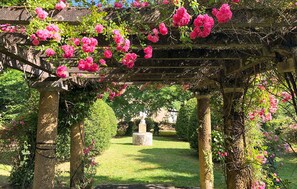 A garden with a pergola covered in pink flowers, stone columns, and a statue in the background. - Awesome home in Ginestet with WiFi (Ginestet)