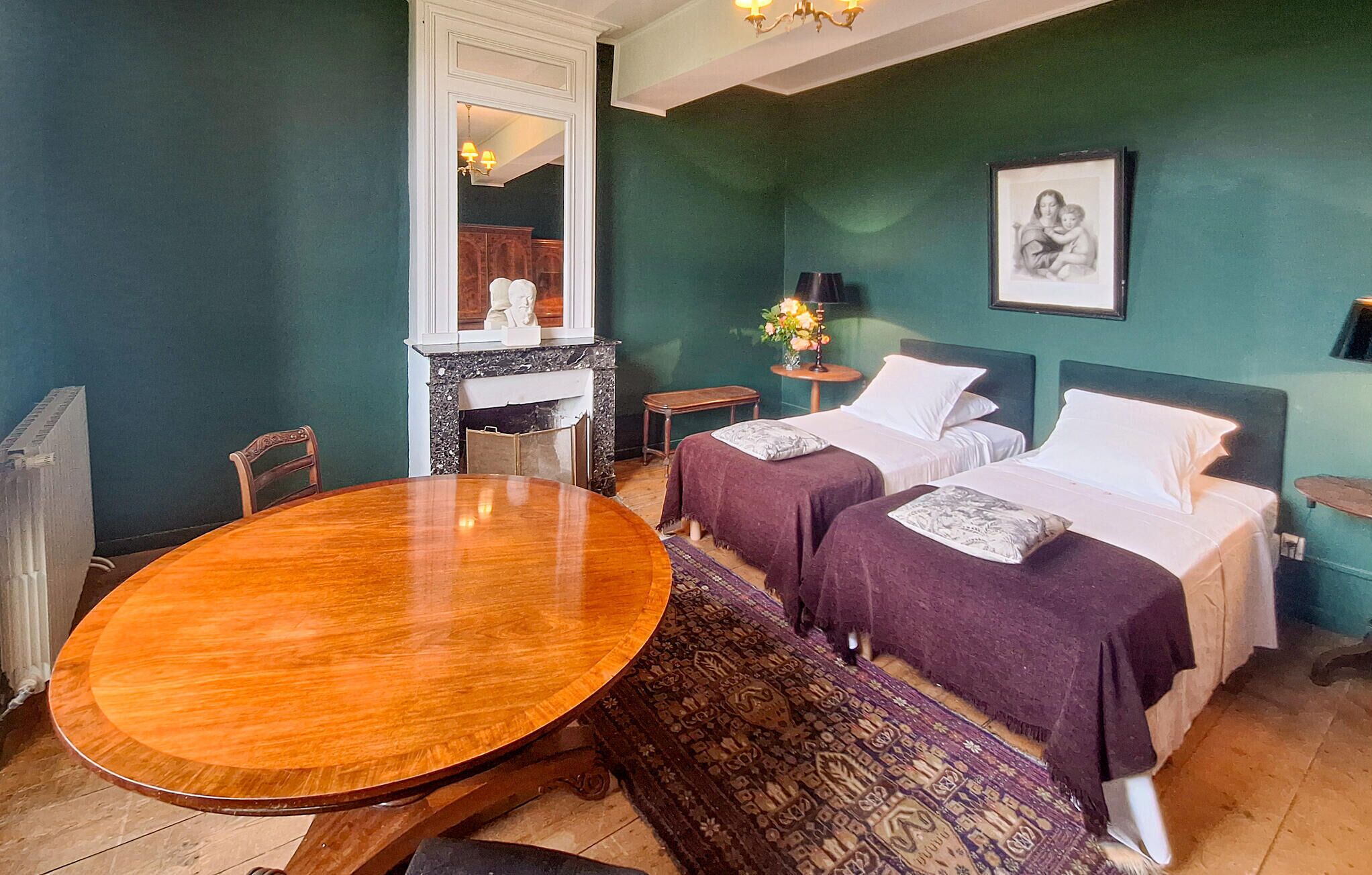 Une pièce avec deux lits, une table ronde en bois, une cheminée et un tableau encadré au mur.