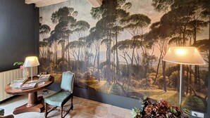 Ein Zimmer mit einer großflächigen Waldmalerei, einem runden Holztisch mit Zeitschriften, einem grünen Polsterstuhl und einer Stehlampe.