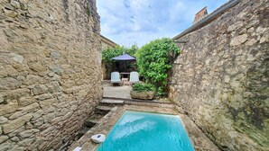 Ein Poolbereich mit Steinwänden, einem kleinen Pool, zwei gestreiften Liegestühlen und einem Tisch unter einem blauen Sonnenschirm.