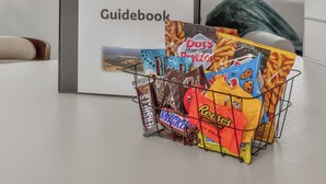 A framed 'House Rules & Guidebook' on a white wall, a snack basket with various chips and chocolates, and a window with blinds.