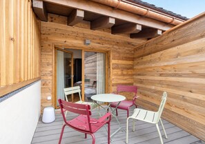 A wooden deck with a table and chairs, a sliding glass door, and a white trash bin. - Luxury Apartment, 1 Bedroom Rahas B Grimentz - By Mrs Miggins (Grimentz)