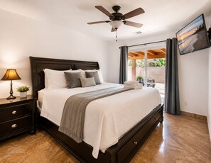 Ein Schlafzimmer mit einem Bett, einem Nachttisch, einer Lampe, einem Deckenventilator und Blick ins Freie.