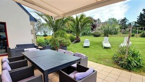 Un patio al aire libre con una mesa, sillas, un área techada y un césped con sillones de descanso.