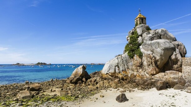 Un paisaje costero rocoso con una pequeña capilla de piedra sobre una formación rocosa, con vistas al mar.