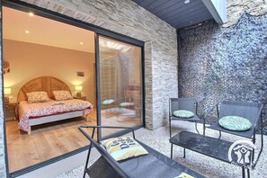 Ein Schlafzimmer mit einem Bett, einer Glastür zu einem Badezimmer sowie einer Sitzecke mit Stühlen und Kissen.