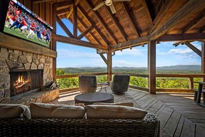 Eine Holzterrasse mit Korbmöbeln, einem Steinkamin und einem Fernseher, der ein Sportspiel zeigt.