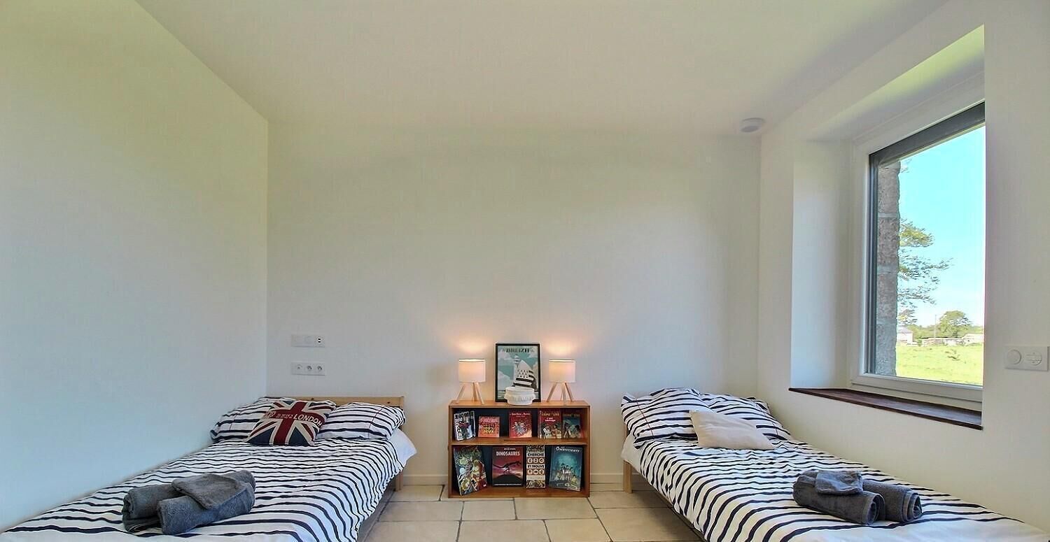 Une petite pièce bien éclairée, avec deux lits, une table de chevet avec des livres, une fenêtre et une vue sur l’extérieur.