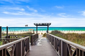 Ein hölzerner Steg führt zu einem Strand mit einer Pergola und einer Flagge.