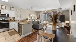 Una cuina moderna amb una illa central, una zona de dinar amb taula i cadires, un sofà i una finestra gran amb cortines.