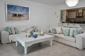 Ein Wohnzimmer mit einer weißen Sofagarnitur, einem Couchtisch mit Dekoration und einem Bild von einer Strandlandschaft.