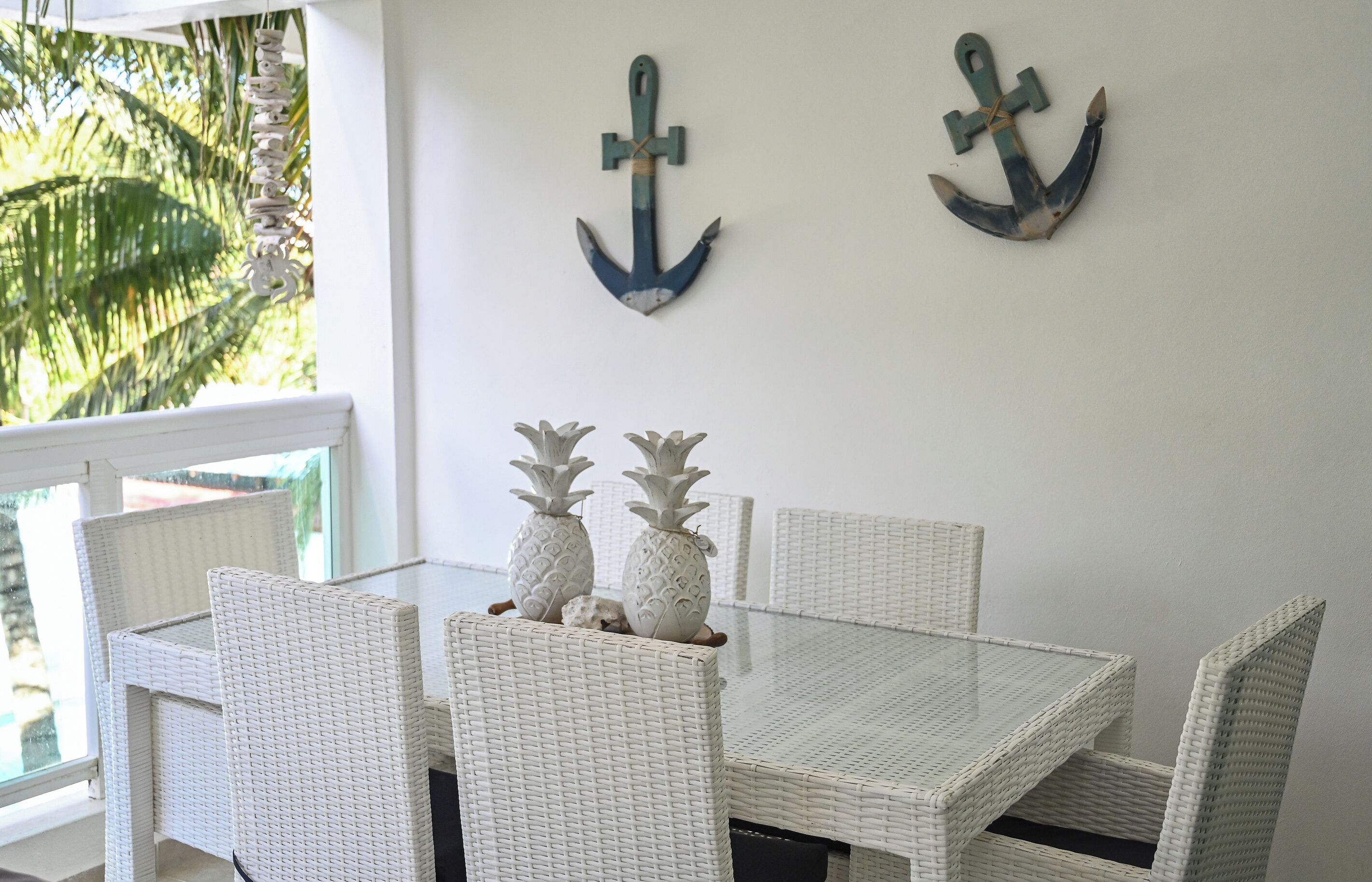 A dining area with white wicker furniture, two anchors on the wall, and a view of palm trees outside.