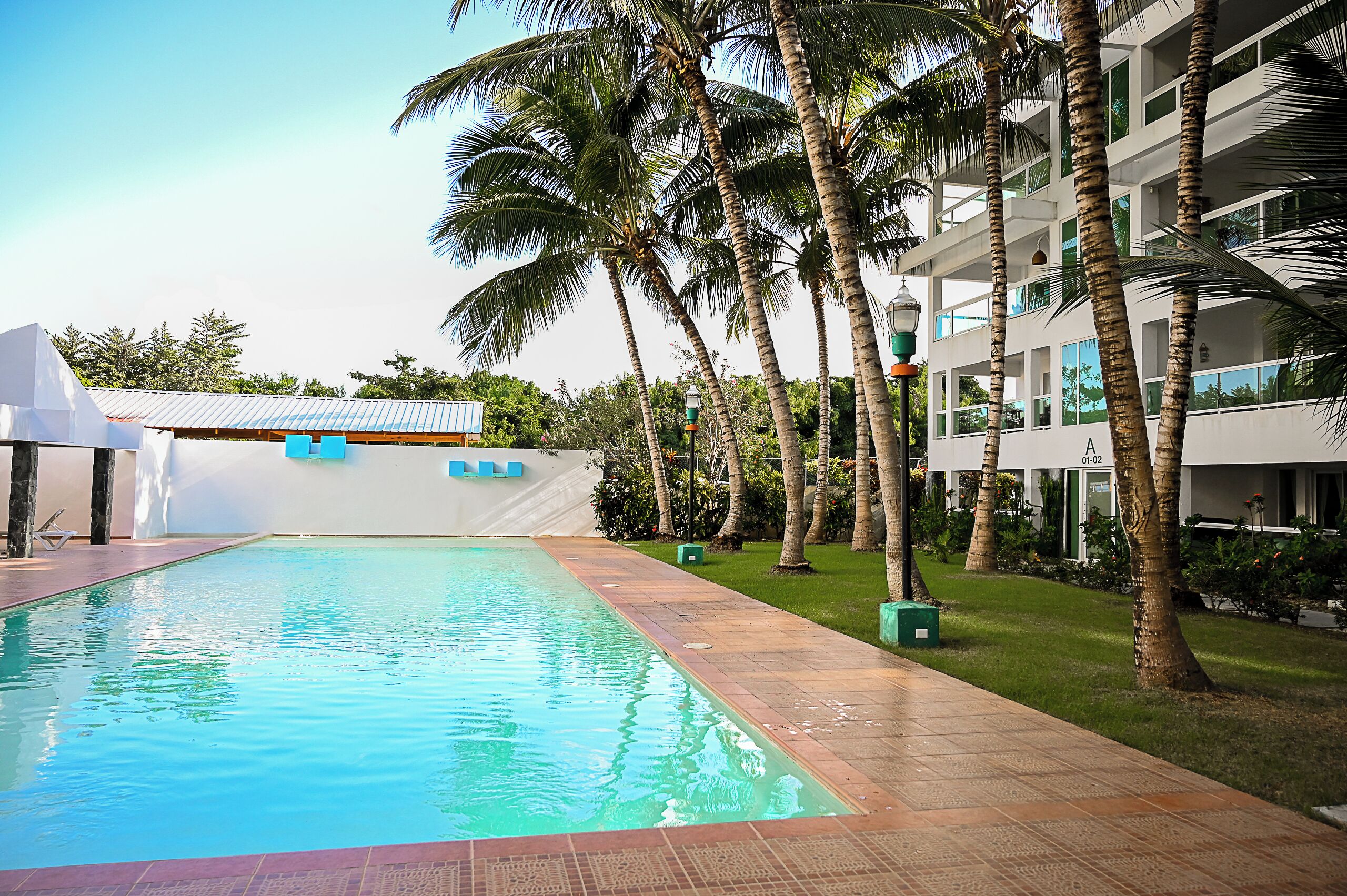 A swimming pool with a tiled deck, surrounded by palm trees and a building with balconies.