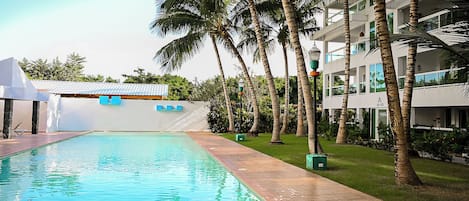 A swimming pool with a tiled deck, surrounded by palm trees and a building with balconies.