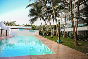 A swimming pool with a tiled deck, surrounded by palm trees and a building with balconies. - Modern 2-Bedroom Apartment in Bibijagua | Enjoy the Beach Just Steps Away (Playa Bibijagua)