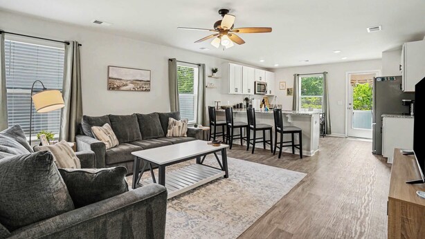 A modern living room with a sofa, coffee table, and ceiling fan. There is a kitchen area with a dining table and chairs. - Whispering Waves in OIB! (Ocean Isle Beach)