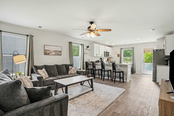 A modern living room with a sofa, coffee table, and ceiling fan. There is a kitchen area with a dining table and chairs. - Whispering Waves in OIB! (Ocean Isle Beach)
