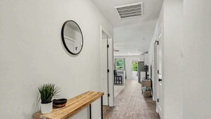 A hallway with a wooden console table, a potted plant, a round mirror, and a ceiling vent. - Whispering Waves in OIB! (Ocean Isle Beach)