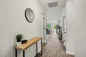 A hallway with a wooden console table, a potted plant, a round mirror, and a ceiling vent. - Whispering Waves in OIB! (Ocean Isle Beach)