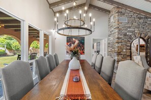 A dining area with a long wooden table, grey upholstered chairs, a chandelier, and a stone fireplace. - Desert Rose - Historic Pink Hacienda with Heated Pool & Billiards (Tempe)