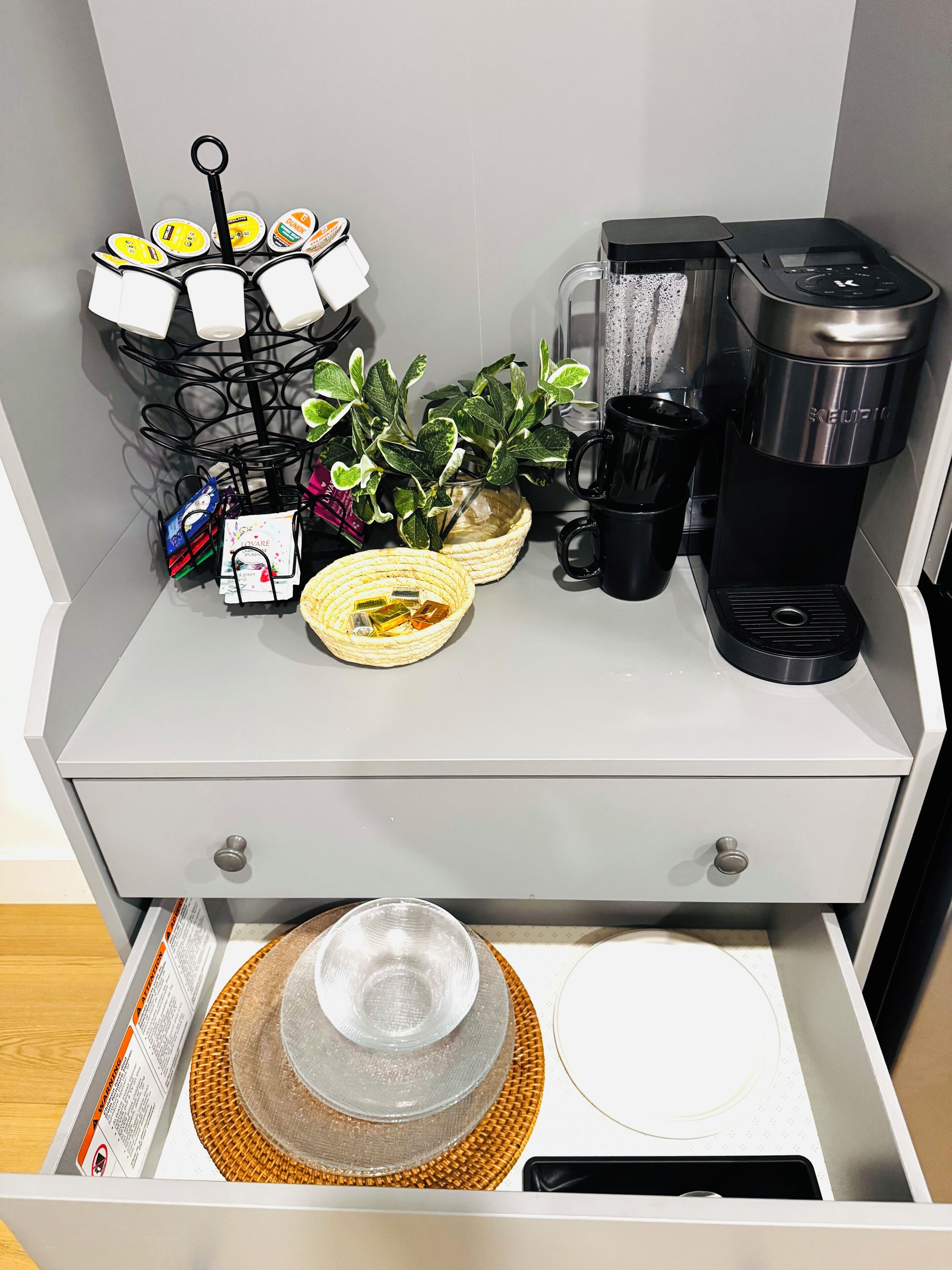 A kitchen counter with a coffee maker, a plant, a basket of tea bags, and a coffee machine.