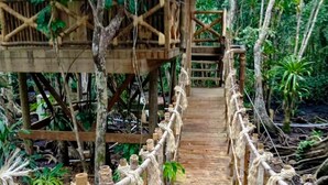 Eine Holzbrücke mit Seilgeländern, die zu einem Gebäude mit einem Strohdach führt.