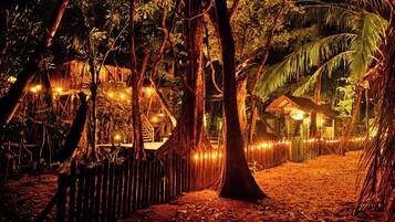 A forested area with a wooden structure and a thatched-roof building, surrounded by trees and illuminated by string lights.