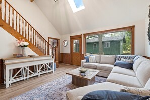 A living room with a wooden staircase, a sofa, a coffee table, and a view of a green house through a large window. - Northlight Lodge | Dual-Lake Sanctuary (Traverse City)