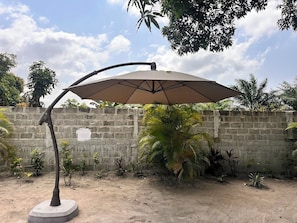 Un patio de concreto con una sombrilla grande, rodeado por un muro de piedra bajo y diversas plantas.