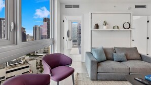 A modern living room with a grey sofa, purple chairs, and a city view through large windows. - Premium 2BR Condo | Beautiful City View (Miami)