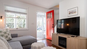 Een woonkamer met een hoekbank, een televisie op een houten kast, een rode deur en een raam met jaloezieën.
