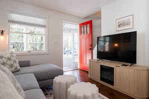 Un salon avec un canapé d’angle, un téléviseur posé sur un meuble en bois, une porte rouge et une fenêtre avec des stores.